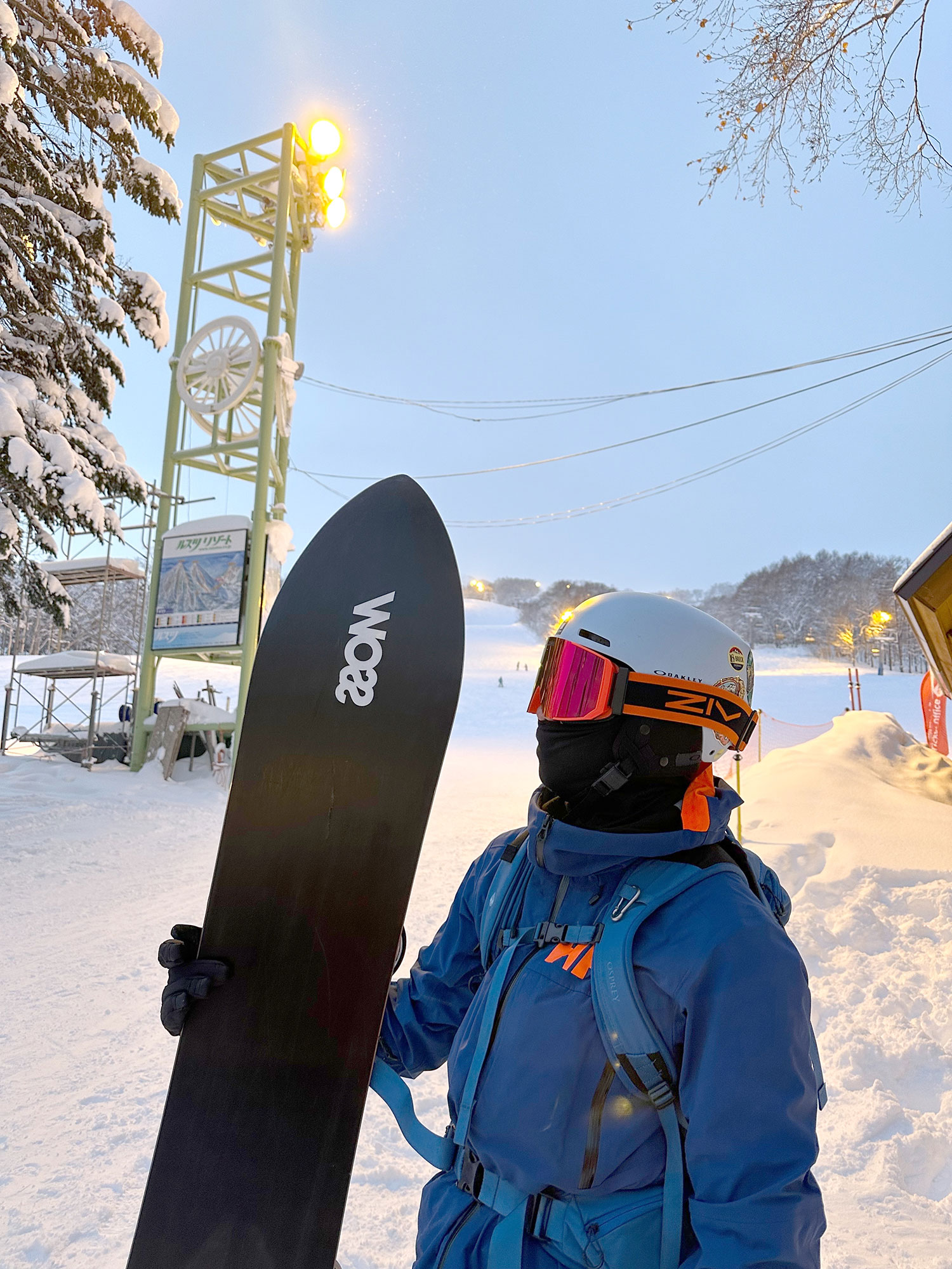 戴著橘色ZIV雪鏡的滑雪者，穿著藍色的防寒大衣，手拿滑雪板