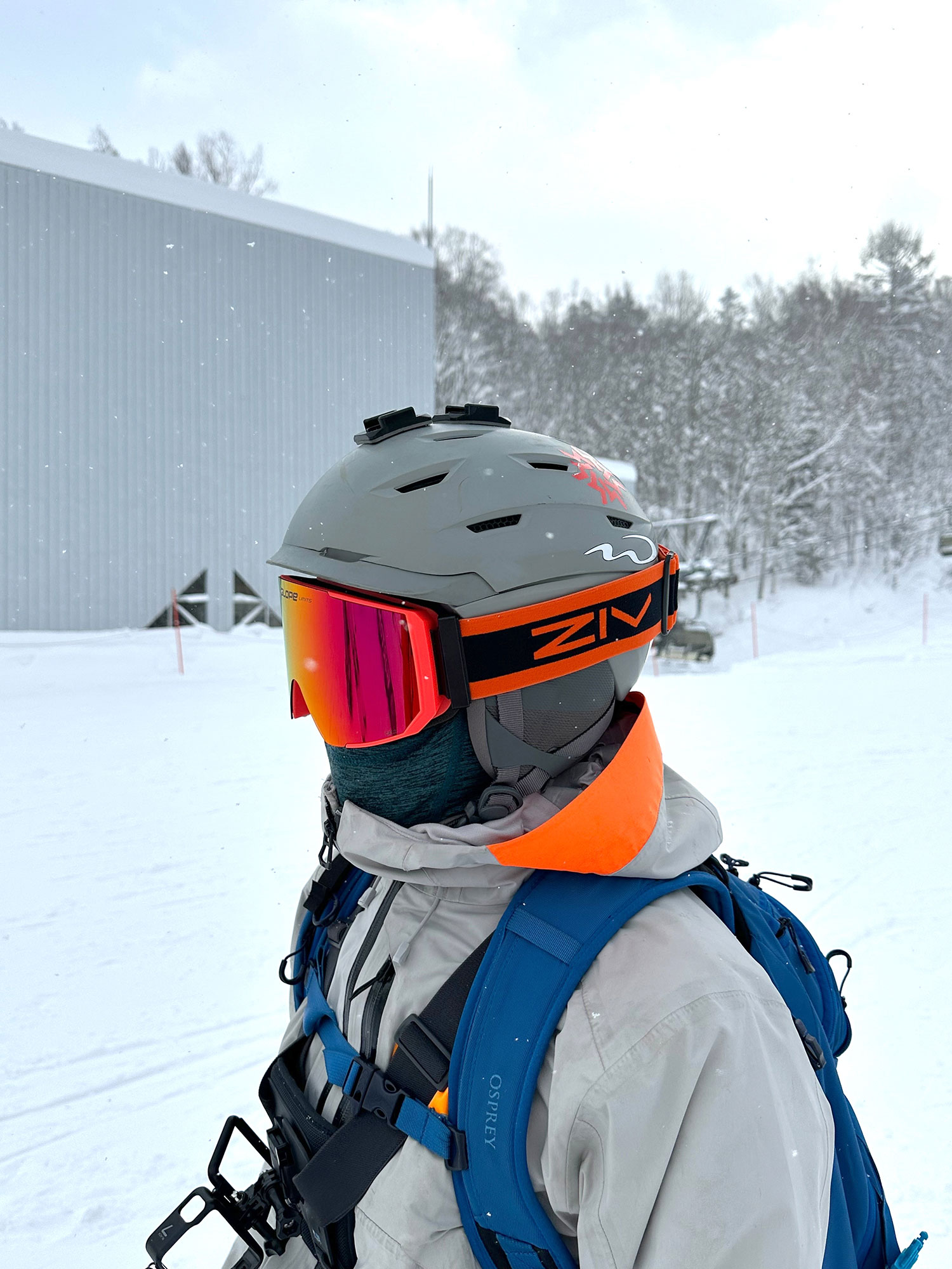 A skier wearing orange ZIV snow goggles, dressed in light gray cold-resistant clothing, carrying a blue backpack.