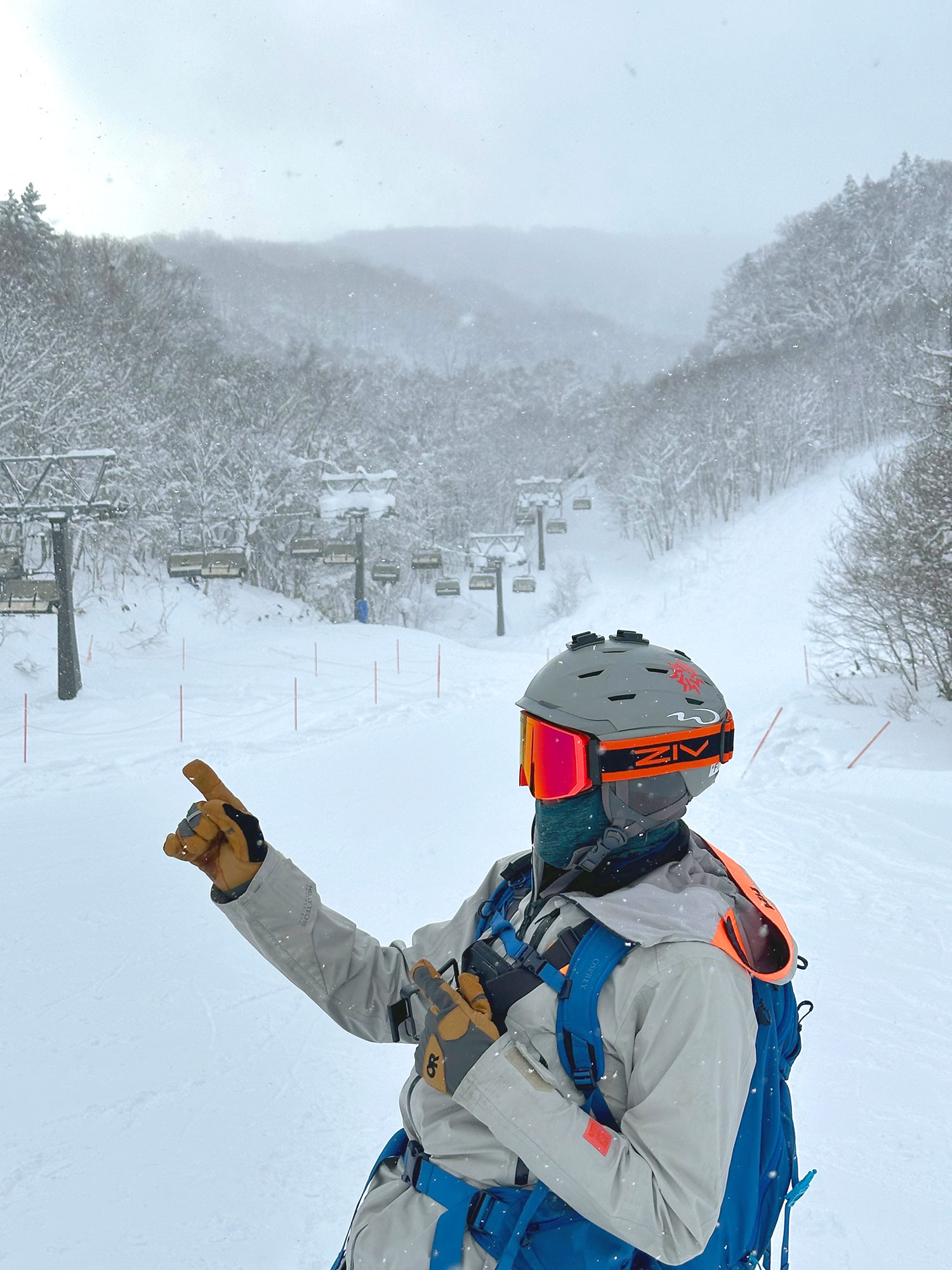 A skier wearing orange ZIV snow goggles, dressed in light gray cold-resistant clothing, carrying a blue backpack, and wearing mustard-yellow cold-resistant gloves.