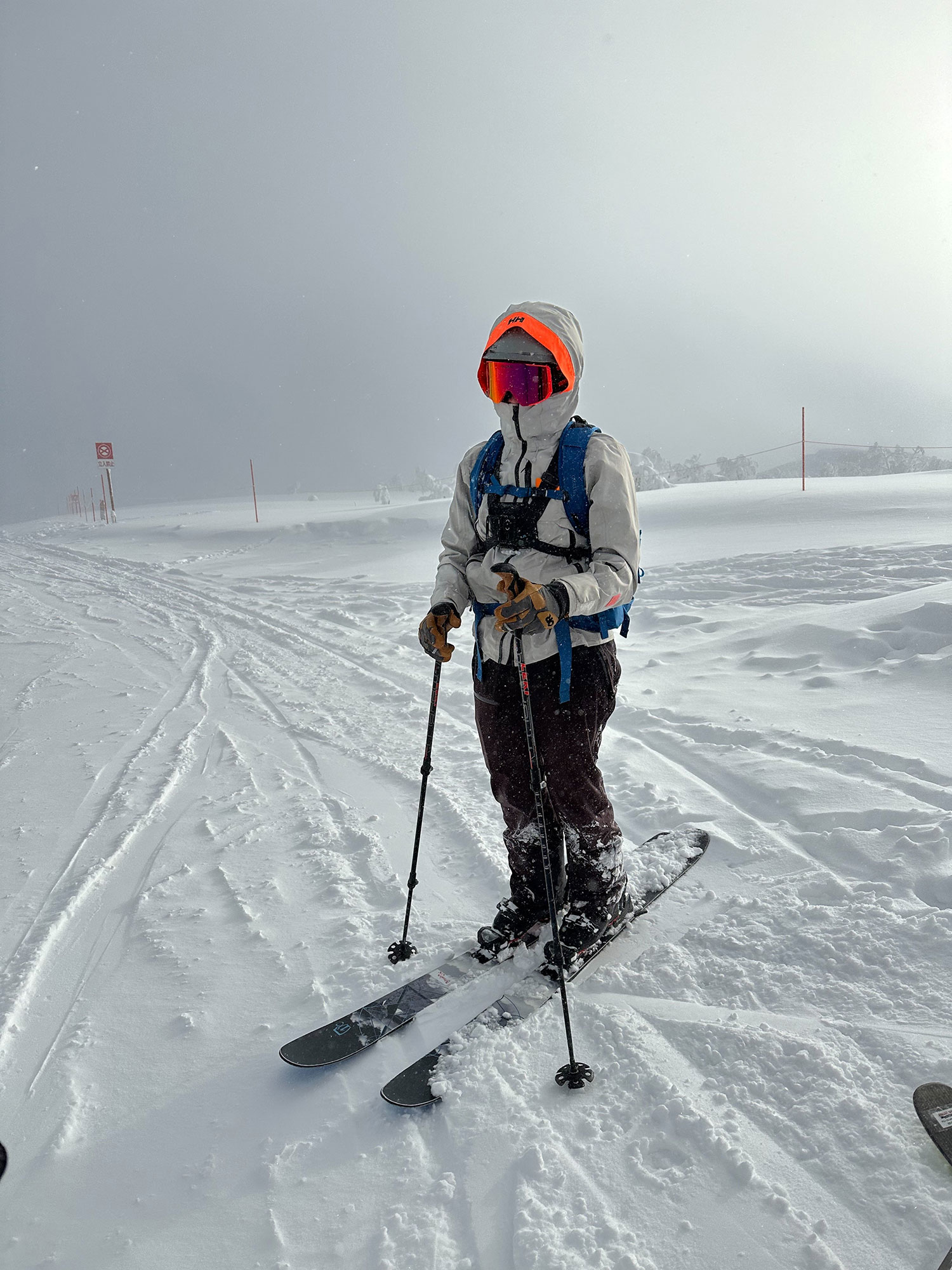 A skier wearing orange ZIV snow goggles, holding ski poles, and wearing snowboard boots.